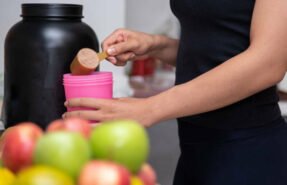 Healthy women preparing a whey protein after doing weight training in the kitchen with fresh fruits as a blurred foreground.